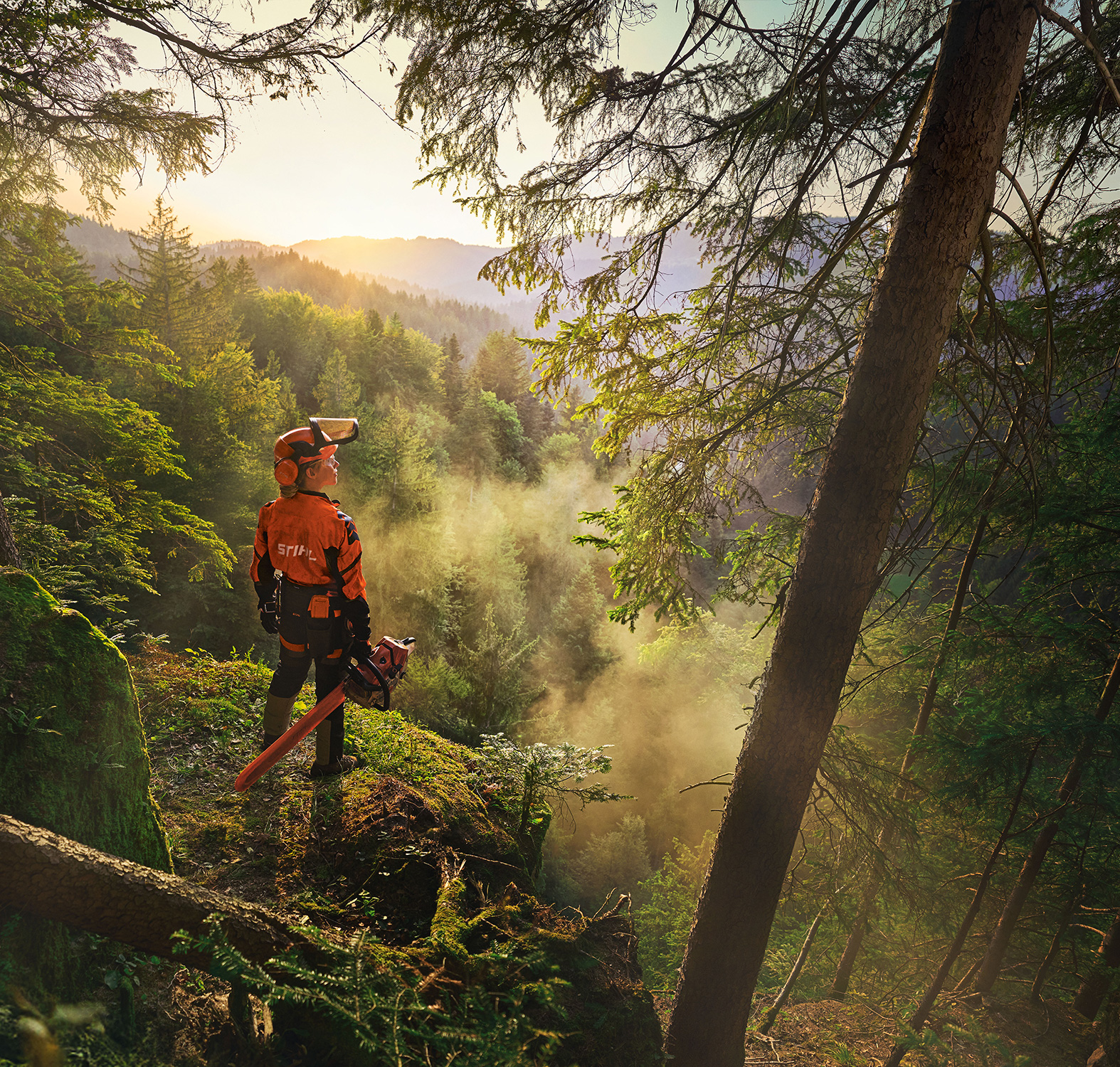 A woman is standing at the edge of a cliff in a saturated green forest. She is wearing orange STIHL workwear, the ADVANCE ProCom Headset, and holding a STIHL MS Chainsaw. 