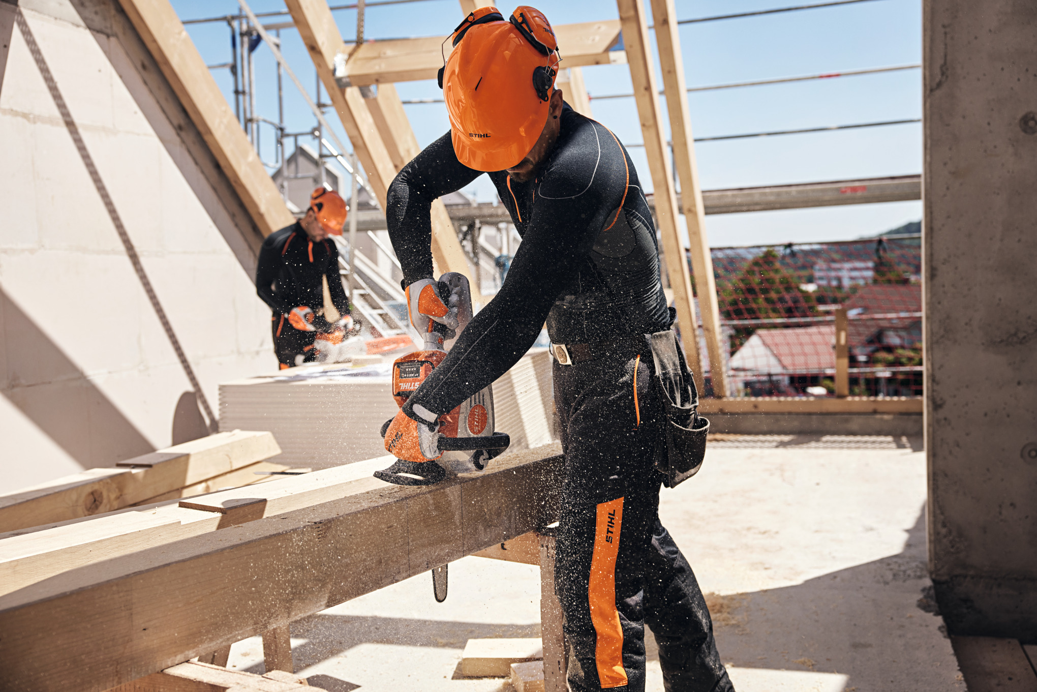A STIHL professional in a loft saws a wooden beam with a cordless chainsaw, while another worker uses a cordless chainsaw in the background