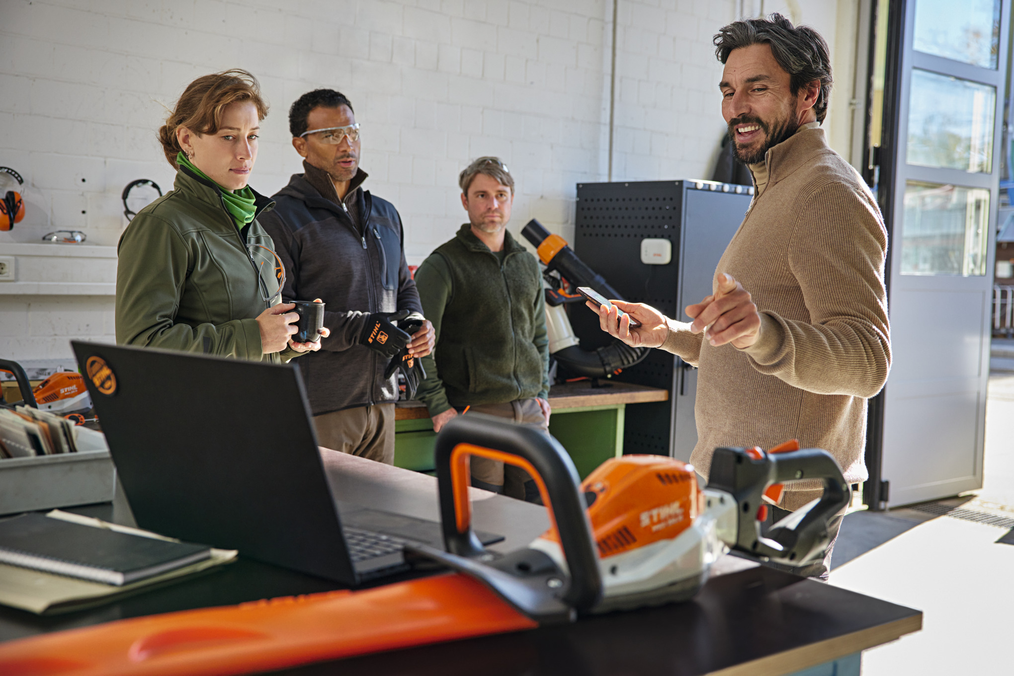 Four people standing in a workshop as one person explains the STIHL connected App on a smartphone