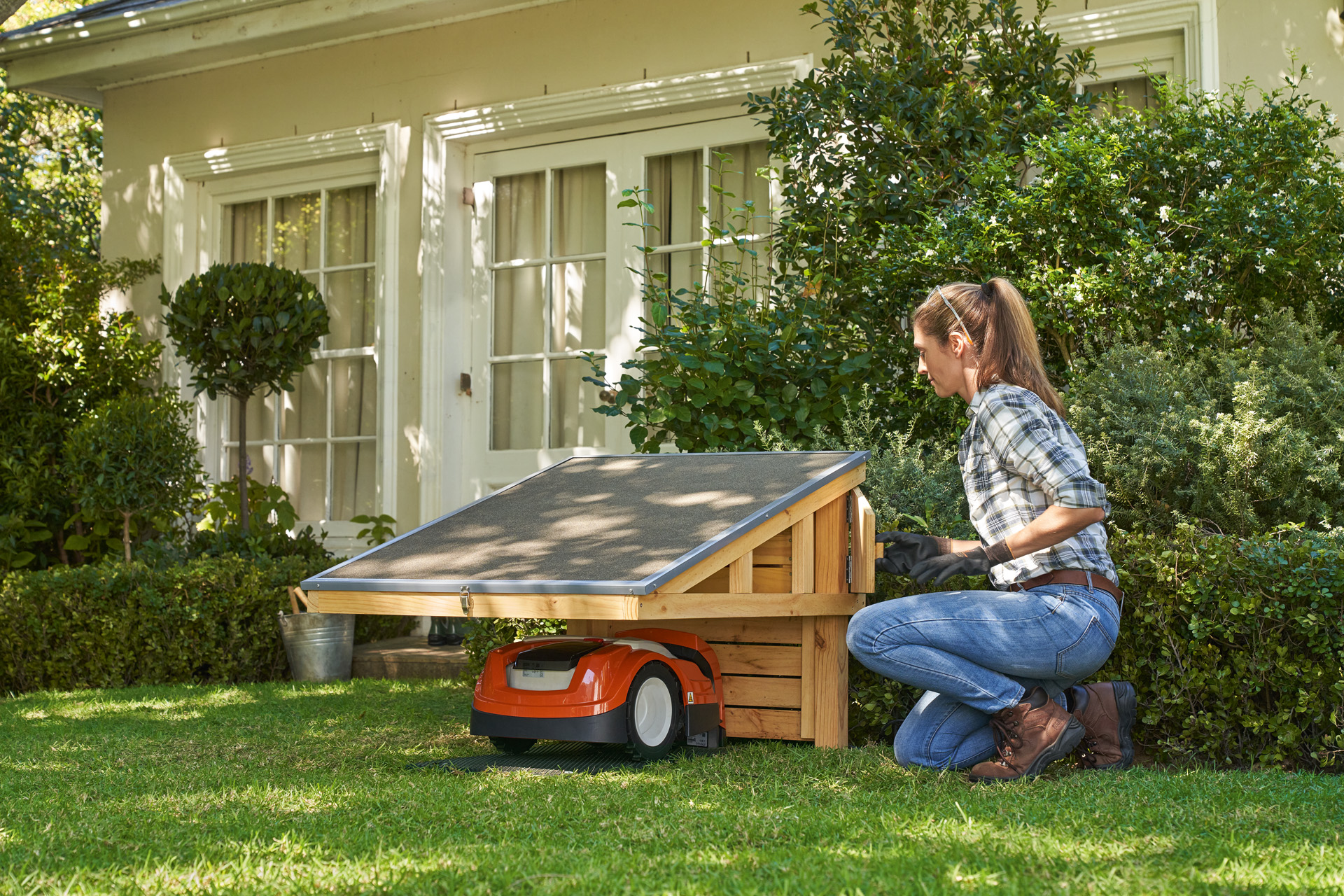Woman wearing work gloves kneeling on grass next to a STIHL iMOW® robot mower inside a DIY garage. 