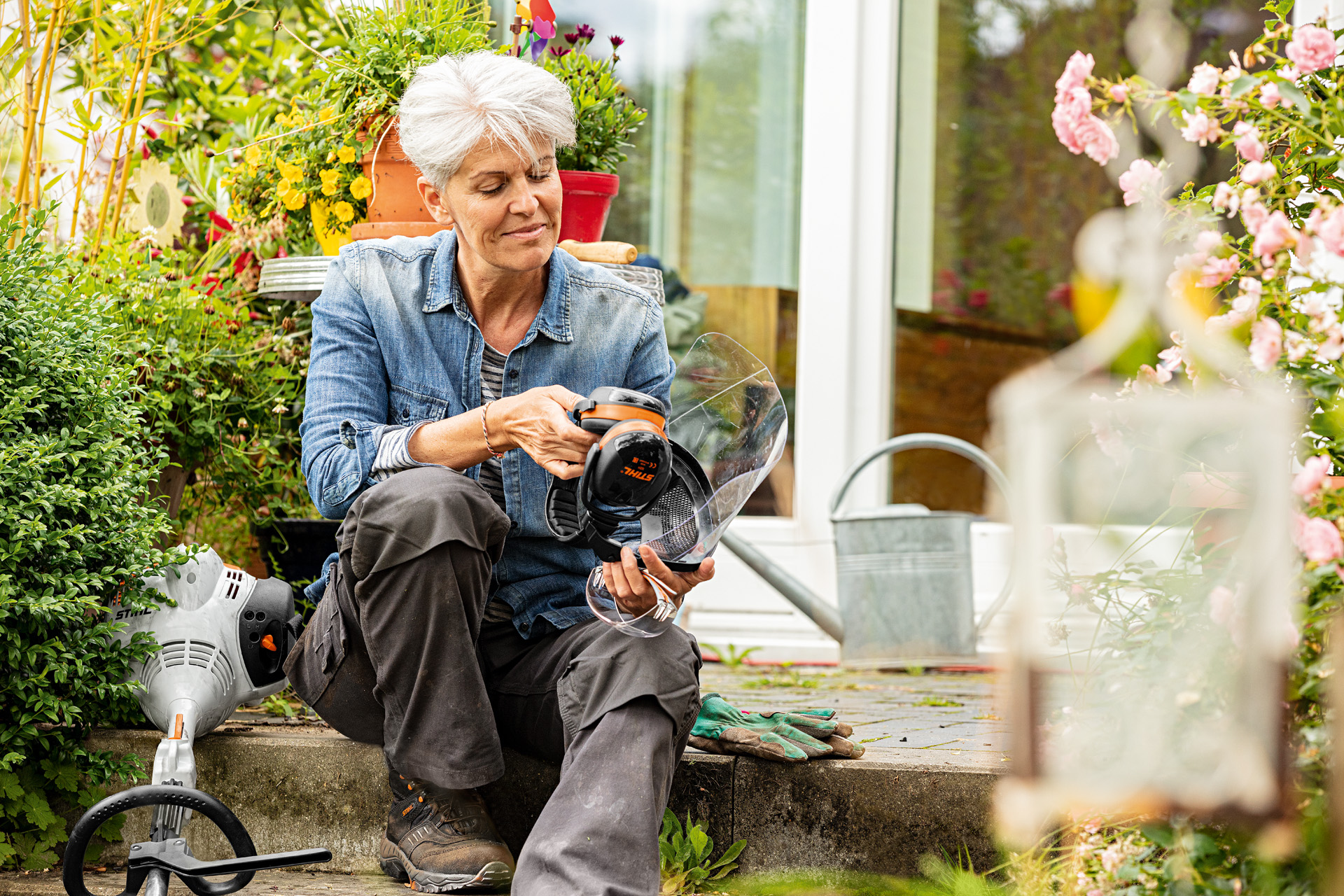 A woman sitting among blooming plants next to a STIHL FS 38 brushcutter and looking at some protective equipment