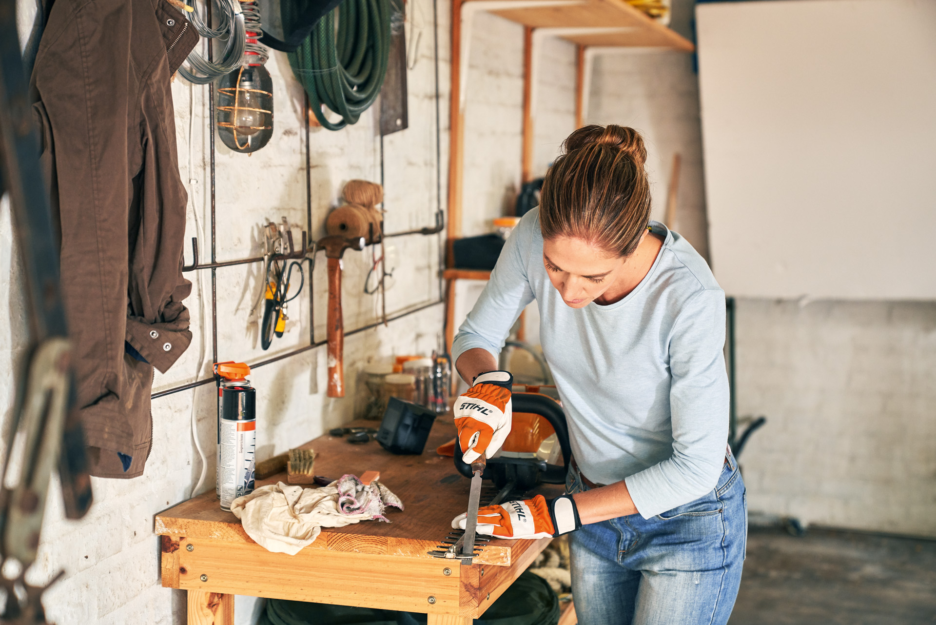 A woman wearing protective gloves using a flat file and sharpening a hedge trimmer on a wooden workbench