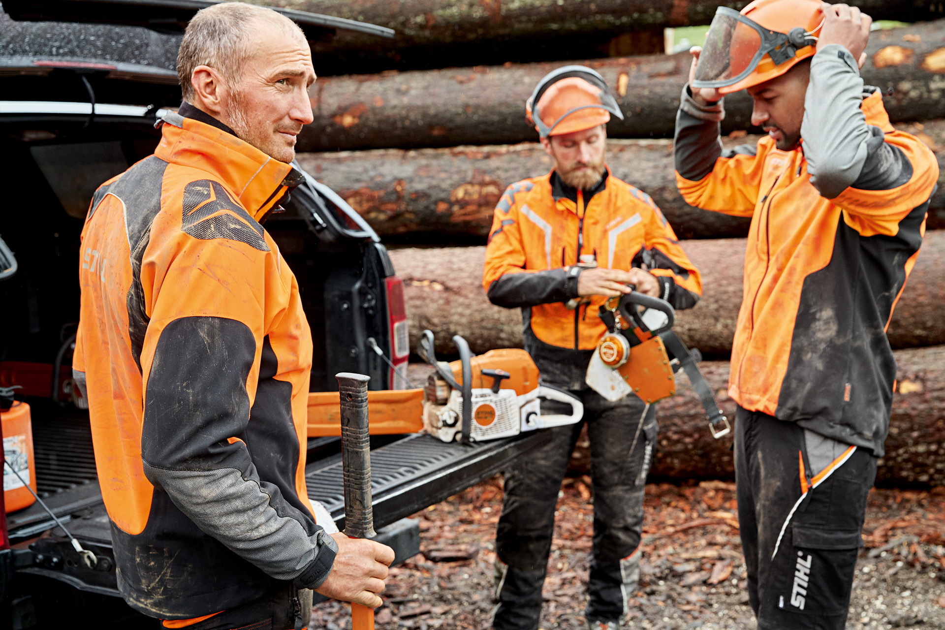 Three men wearing STIHL protective clothing, getting ready to use a STIHL chainsaw in front of a stack of wood